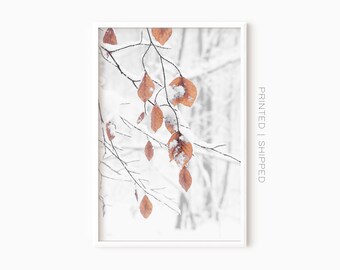 May include: A framed print featuring a winter scene. Brown leaves are dusted with snow and cling to thin branches against a blurred, snowy background. The print is in a white frame, with the text "PRINTED | SHIPPED" on the right side.