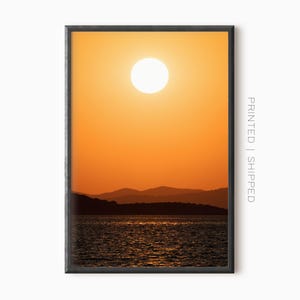 Pode incluir: Impressão emoldurada de um pôr do sol sobre o mar. O sol é um orbe branco brilhante num céu laranja vibrante. Montanhas silhuetadas são visíveis ao longe, e a água reflete o brilho do pôr do sol. O texto "PRINTED | SHIPPED" é visível.