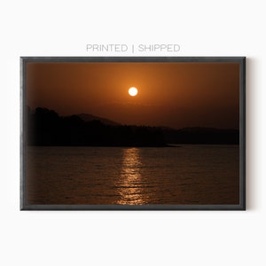 Può includere: Stampa incorniciata di un tramonto sull'acqua. Il sole è un globo arancione brillante, che si riflette sulla superficie dell'acqua. Scure sagome di alberi e colline sono sullo sfondo. Il testo "PRINTED | SHIPPED" è in alto.