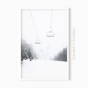 May include: A black and white photograph of a ski lift with empty chairs ascending a snowy mountainside. The trees are covered in snow and the sky is overcast.
