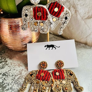 Pode incluir: Um par de brincos de elefante de contas douradas e vermelhas. Os brincos são pendentes e apresentam um elefante de contas douradas e vermelhas com um círculo menor de contas douradas na parte superior.