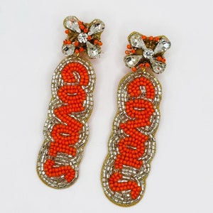 May include: A pair of orange and silver beaded earrings with a flower charm. The earrings spell out the word "LOVE" in orange beads.