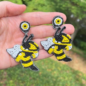 Puede incluir: Un par de pendientes de cuentas con forma de abejas. Las abejas son negras, amarillas y blancas con un amuleto de ojo turco negro y blanco que cuelga de la parte superior.