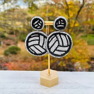 Peut inclure: Une paire de boucles d'oreilles en perles noires et blanches en forme de volley-ball. Les boucles d'oreilles sont présentées sur un support doré.