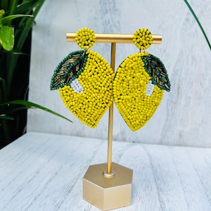 May include: A pair of yellow lemon-shaped beaded earrings with green leaves. The earrings are displayed on a gold stand.