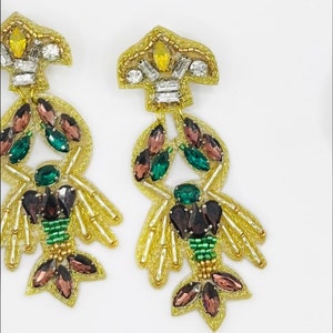 Può includere: Un paio di orecchini dorati con un design a forma di aragosta. Gli orecchini sono decorati con strass verdi, marroni e trasparenti.