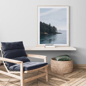 May include: A framed photograph of a scenic landscape with a forested shoreline and a body of water. The water is a light blue color and the sky is a light blue with white clouds. The photo is hanging on a white wall above a wooden shelf.