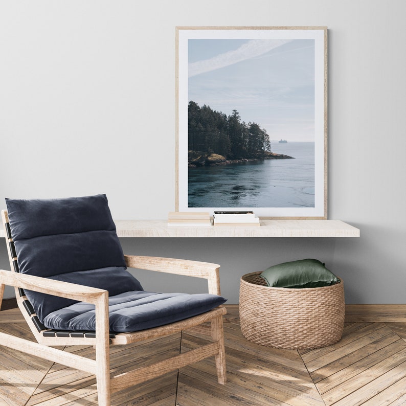 May include: A framed photograph of a scenic landscape with a forested shoreline and a body of water. The water is a light blue color and the sky is a light blue with white clouds. The photo is hanging on a white wall above a wooden shelf.