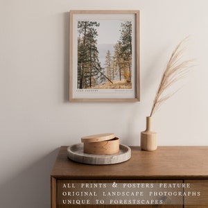 Op de afbeelding: Een ingelijste print van een landschapsfoto met uitzicht op een meer en bomen. De print is in een licht houten lijst en heeft de tekst "Lake Country" en "Okanagan Valley" onderaan gedrukt. De print hangt aan een witte muur.