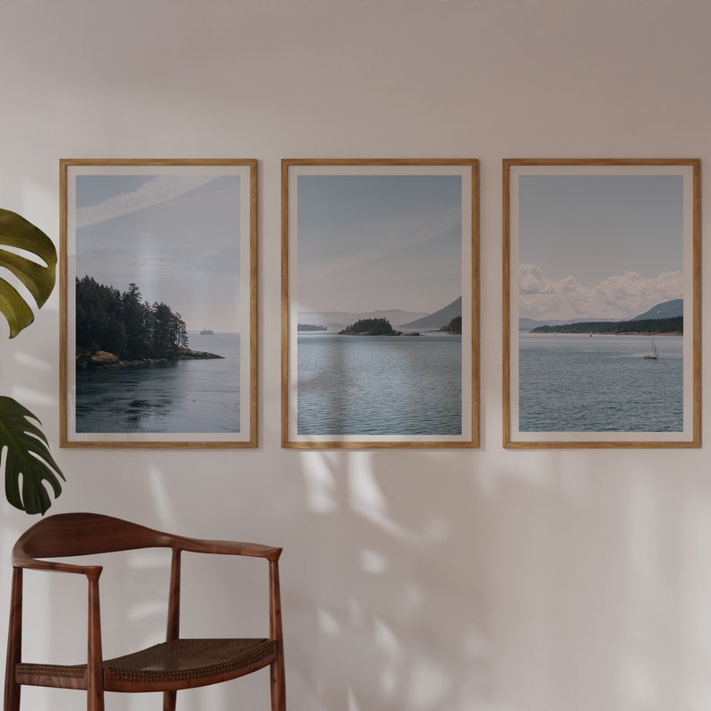 May include: Three framed photographs of a seascape with islands and a sailboat in the distance. The photos are in wooden frames and are hung on a white wall.
