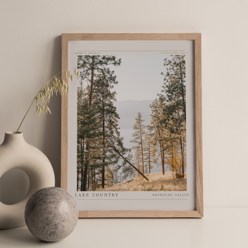 Op de afbeelding: Een ingelijste foto van een boslandschap met hoge dennenbomen en een meer in de verte. De tekst "LAKE COUNTRY" en "OKANAGAN VALLEY" staat onderaan de afbeelding.