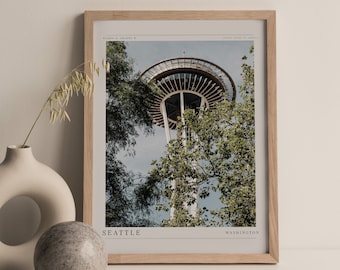 May include: A framed print of the Space Needle in Seattle, Washington. The Space Needle is a tall, slender tower with a circular observation deck at the top. The print is in a light wood frame and has a white background. The text "SEATTLE" and "WASHINGTON" is printed at the bottom of the print.
