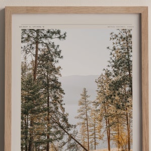 Op de afbeelding: Een ingelijste foto van een boslandschap met hoge dennenbomen op de voorgrond en een wazig uitzicht op een meer op de achtergrond. De afbeelding heeft de titel "50.0537 N, 119.4106 W" en "BRITISH COLUMBIA, CANADA".