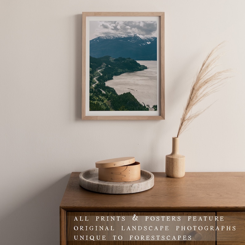 May include: A framed photograph of a mountain range with a winding road leading to a body of water. The photograph is hanging on a white wall above a wooden table with a decorative wooden box and a vase with dried grass.