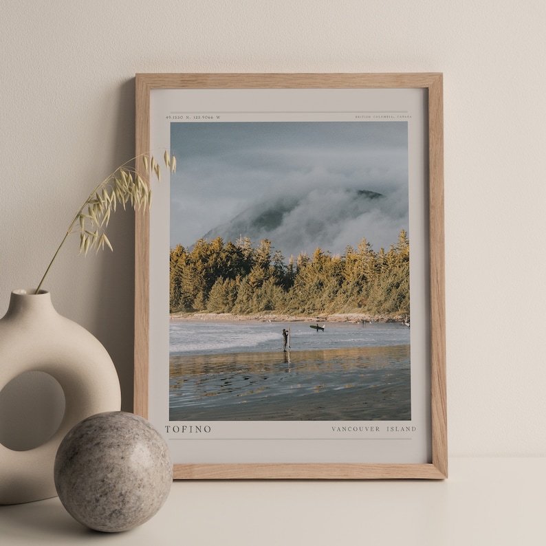May include: A framed print of a beach scene with a mountain in the background. The beach is covered in fog and there are two people surfing in the water. The print has the text "TOFINO" and "VANCOUVER ISLAND" at the bottom.