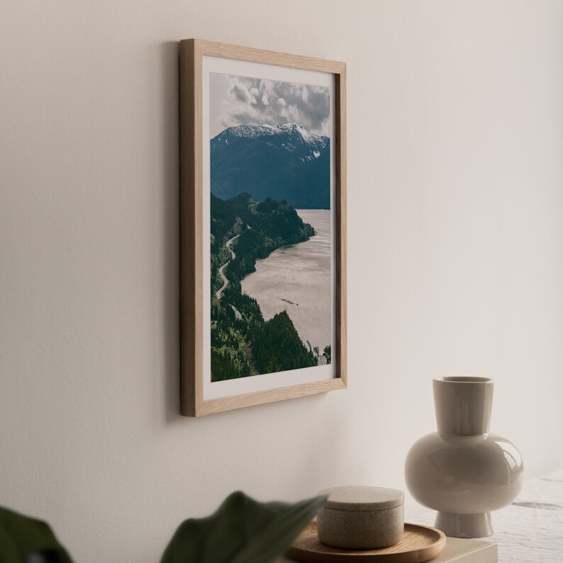May include: A framed photograph of a mountain range with a winding road leading to a body of water. The photo is in a light wood frame and is hanging on a white wall.