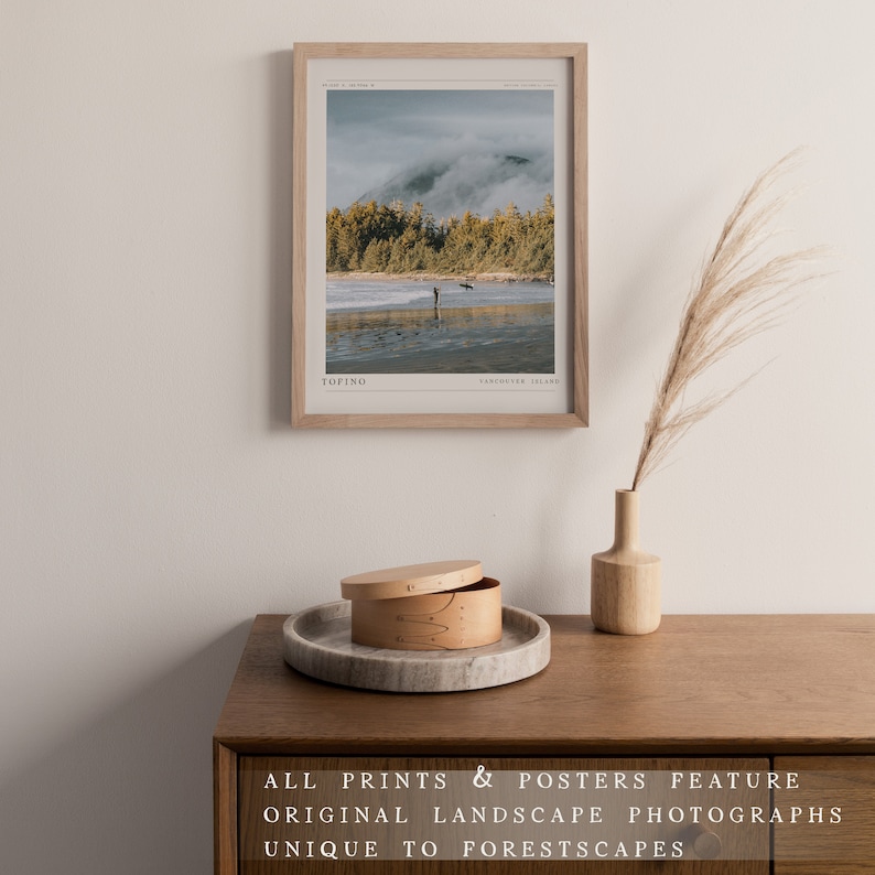 May include: A framed photograph of a beach scene with a person standing in the water. The photo is in a light wood frame and has the text "TOFINO" and "VANCOUVER ISLAND" at the bottom. The photo is hanging on a white wall.