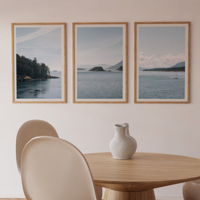 May include: Three framed landscape photographs of a body of water with islands and mountains in the distance. The photos are hung on a white wall above a wooden table with a white ceramic vase.