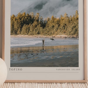 May include: A framed photograph of a surfer standing in the water with a surfboard. The beach is sandy and the water is calm. The background is a forest with a cloudy sky. The text "TOFINO" and "VANCOUVER ISLAND" is printed at the bottom of the image.