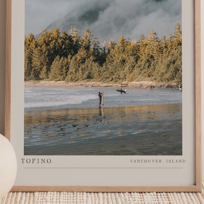 May include: A framed photograph of a surfer standing in the water with a surfboard. The beach is sandy and the water is calm. The background is a forest with a cloudy sky. The text "TOFINO" and "VANCOUVER ISLAND" is printed at the bottom of the image.