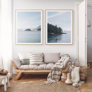 May include: Two framed landscape photographs of a body of water with islands and trees in the distance. The photographs are hanging on a white wall above a wooden futon with a throw blanket and pillows.