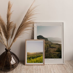 Puede incluir: Dos fotograf&iacute;as de paisajes enmarcadas de un valle verde con monta&ntilde;as en el fondo. La foto superior est&aacute; en un marco blanco y la foto inferior est&aacute; en un marco de madera natural. Ambas fotos est&aacute;n sobre una superficie tejida marr&oacute;n claro.