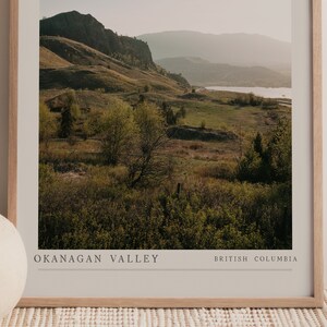 Puede incluir: Una fotograf&iacute;a enmarcada de un paisaje esc&eacute;nico con colinas onduladas, campos verdes y un lago en la distancia. El texto "OKANAGAN VALLEY BRITISH COLUMBIA" est&aacute; impreso en la parte inferior de la imagen.