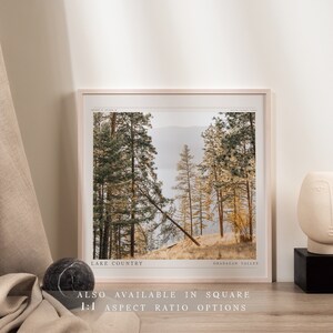 Op de afbeelding: Een ingelijste foto van een boslandschap met hoge bomen en een meer in de verte. De afbeelding heeft de titel "Lake Country" en "Okanagan Valley". De tekst "Also available in square 1:1 aspect ratio options" staat onder de afbeelding.