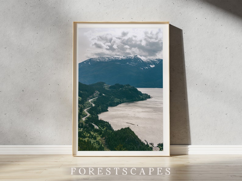 May include: A framed photograph of a winding road leading to a mountain range with snow-capped peaks. The road is surrounded by lush green trees and a body of water. The sky is cloudy and the overall tone of the image is serene.