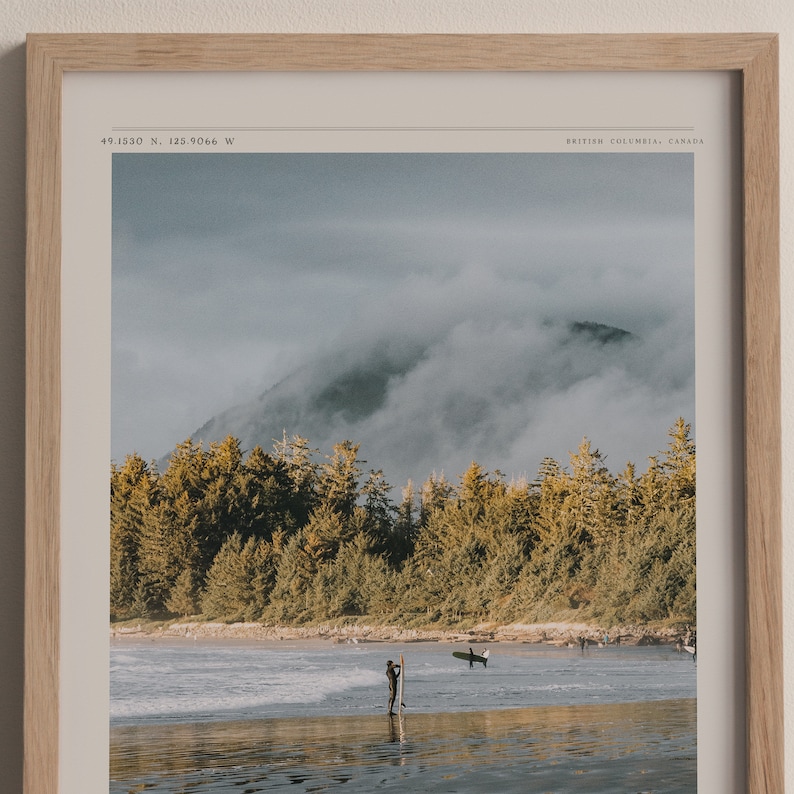 May include: A framed photograph of a misty mountain landscape with a beach in the foreground. The beach is empty except for a lone surfer standing in the water with a surfboard. The text "49.1530 N, 125.9066 W" and "BRITISH COLUMBIA, CANADA" are printed in the top corners of the frame.