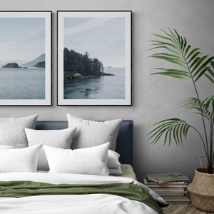 May include: Two framed landscape photographs of a forested island with a body of water in the foreground. The photographs are hanging on a gray wall above a white bed with green bedding and white pillows. A potted plant is on the right side of the image.