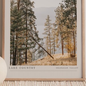 Op de afbeelding: Een ingelijste foto van een boslandschap met hoge dennenbomen en een meer in de verte. De tekst "LAKE COUNTRY OKANAGAN VALLEY" staat onderaan de afbeelding.