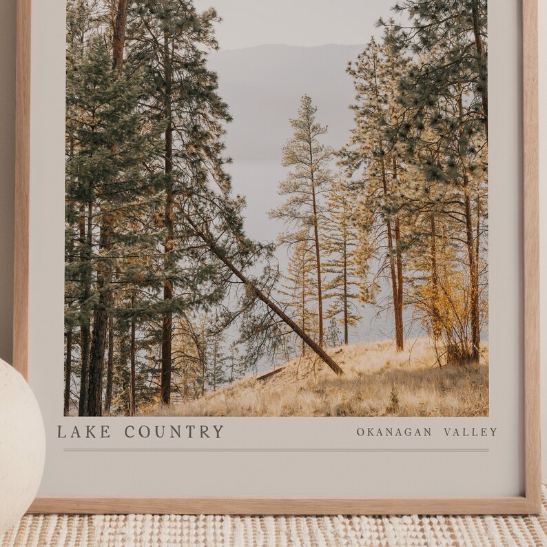 Op de afbeelding: Een ingelijste foto van een boslandschap met hoge dennenbomen en een meer in de verte. De tekst "LAKE COUNTRY OKANAGAN VALLEY" staat onderaan de afbeelding.