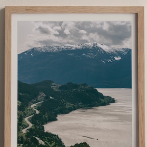 May include: A scenic photograph of a winding road leading through a lush green forest along a body of water. The water is calm and reflects the cloudy sky. In the distance, a snow-capped mountain range rises above the trees.