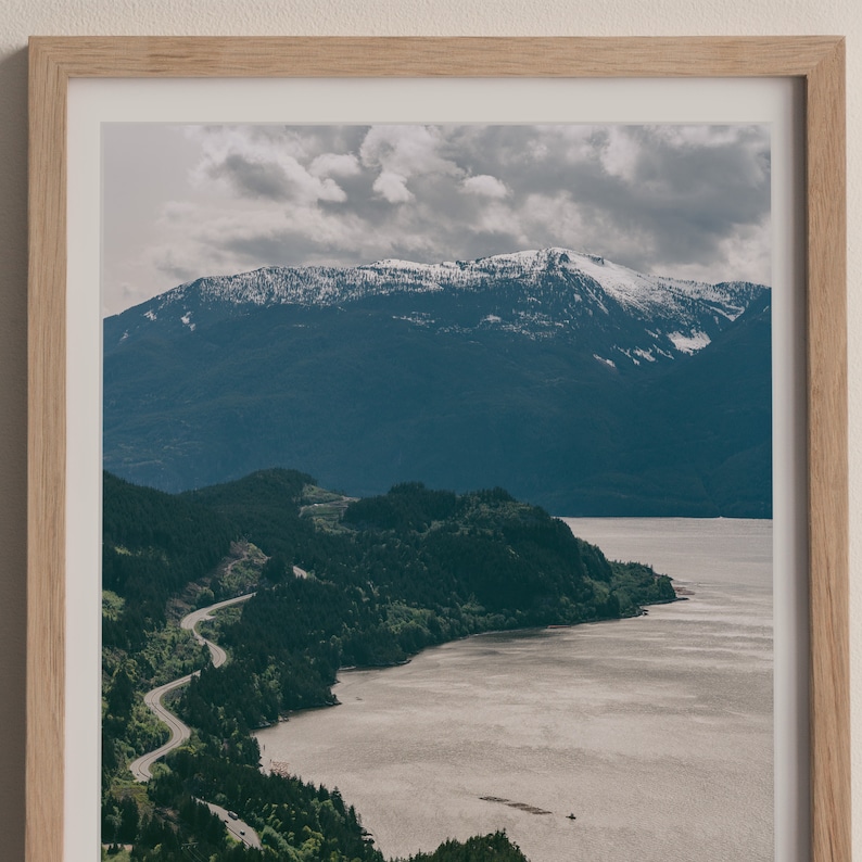 May include: A scenic photograph of a winding road leading through a lush green forest along a body of water. The water is calm and reflects the cloudy sky. In the distance, a snow-capped mountain range rises above the trees.