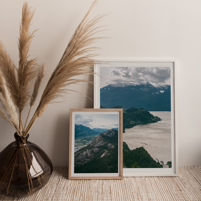 May include: Two framed landscape photographs of mountains and water. The larger frame is white and the smaller frame is wood. The frames are on a light brown surface.