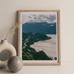May include: A framed photograph of a winding road along a coastline with a mountain range in the background. The sky is cloudy and the water is a light brown color.