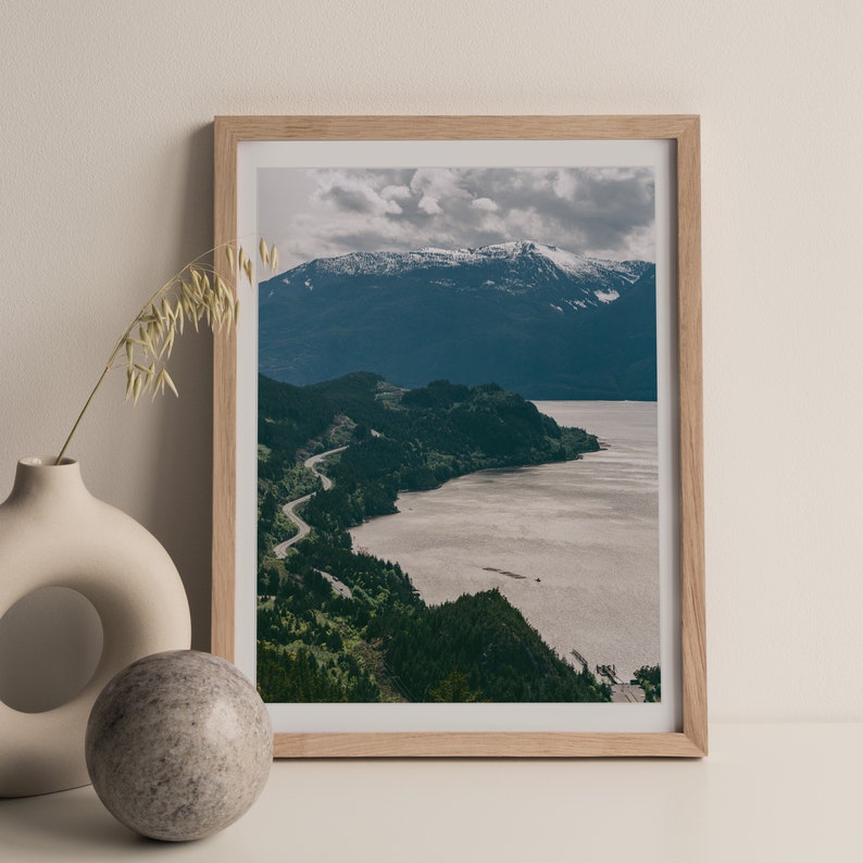May include: A framed photograph of a winding road along a coastline with a mountain range in the background. The sky is cloudy and the water is a light brown color.