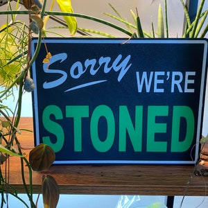 Könnte beinhalten: Ein schwarzes und grünes Schild mit dem Text "Sorry We're Stoned" in weißen Buchstaben. Das Schild hängt an einem Holzregal mit grünen Pflanzen im Hintergrund.