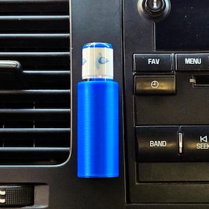 May include: A blue car air freshener with a white and blue top, attached to a car's air vent. The air freshener is cylindrical and has a simple design. The car's dashboard features buttons for radio and menu functions.