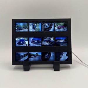 May include: A black rectangular digital display frame featuring a grid of twelve individual screens. Each screen shows a different photograph of cars, including close-ups of wheels, rear views, and exterior shots, connected by a white power cord.