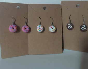 May include: Three pairs of dangle earrings featuring miniature donut charms. One pair is pink with colorful sprinkles, one pair is white with colorful sprinkles, and one pair is brown with white frosting.
