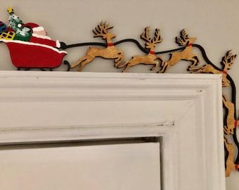 May include: A wooden door frame topper featuring a painted Santa Claus in a red sleigh filled with colorful gifts and a Christmas tree, pulled by a team of tan reindeer with red collars, connected by a black metal wire along the top and side.