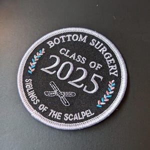 May include: Circular black embroidered patch with white text: "BOTTOM SURGERY CLASS OF 2025 SIBLINGS OF THE SCALPEL." The patch has a white border and a design of crossed surgical tools. Pink and blue accents are on the border.