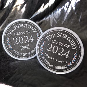 May include: Two black and white embroidered patches with white stitching. The left patch has the text "ORCHIEСTOМУ CLASS OF 2024 SISTERS OF THE SCALD" and a pair of crossed scissors. The right patch has the text "TOP SURGERY CLASS OF 2024 BROTHERS UNBOUND" and a series of stitches.