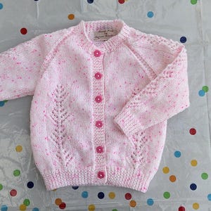 May include: A pale pink, hand-knitted cardigan with a speckled design. The sweater has a round neck, long sleeves, and pink buttons. Decorative leaf patterns are knitted into the front. The garment is made of soft yarn.