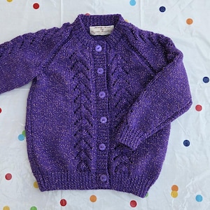 Peut inclure: Un cardigan tricoté violet avec un col rond et des manches longues. Le cardigan a une fermeture boutonnée sur le devant et un motif texturé. Les manches ont un motif en tricot torsadé. Le vêtement est en matière douce, peut-être en laine.