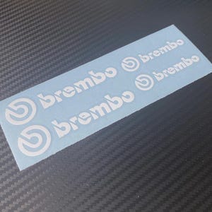May include: White Brembo decals on a light blue backing. The decals feature the Brembo logo and the word "brembo" in a stylized font. The decals are rectangular and are likely for automotive use.