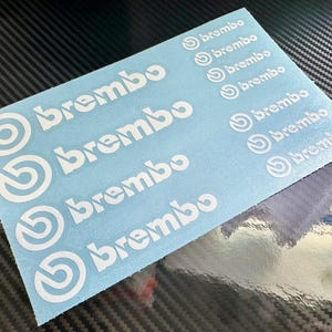 May include: A light blue rectangular sheet featuring multiple white Brembo brand logos and text. The sheet contains four large logos on the left side and seven smaller logos arranged in a column on the right side, all printed in a clean white font.