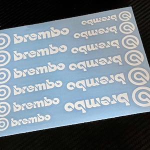 May include: A sheet of white Brembo decals on a light blue background. The decals are arranged in rows and columns, with the Brembo logo and text in a clean, sans-serif font. The background is a solid, light blue color.